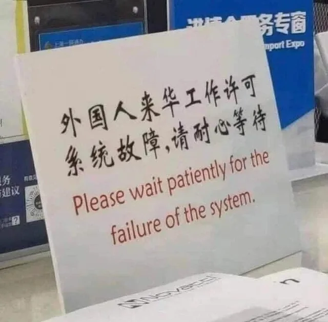 Photo of a white sign on a counter with Chinese characters above red English text reading "Please wait patiently for the failure of the system." A blue banner behind it partly reads "nport Expo". A paper in the foreground reads "Novatel".
