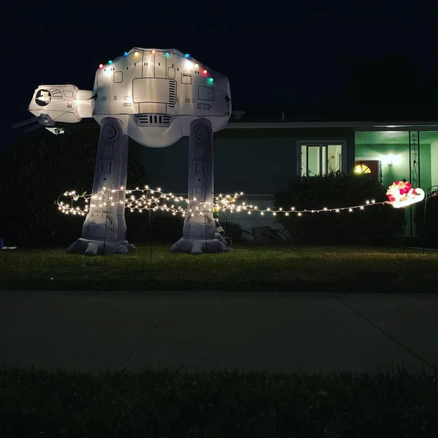 A nighttime front yard with a giant inflatable Star Wars AT-AT walker strung with colorful Christmas lights. A long strand of white lights trails from its head like a tow cable to a glowing Santa in a sleigh near the house porch. No text.
