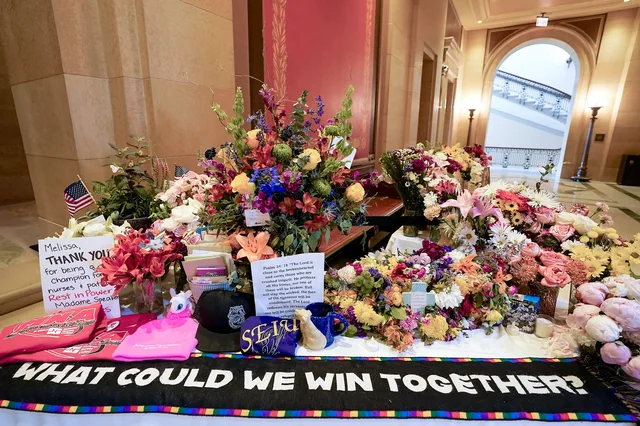 A memorial table of bouquets, cards and keepsakes in a marble hall with an arched doorway. A note reads "Melissa, THANK YOU for being a champion for nurses & patients. Rest in Power Madame Speaker." A printed page quotes Psalm 34:18. A black banner with a rainbow border reads "WHAT COULD WE WIN TOGETHER?"