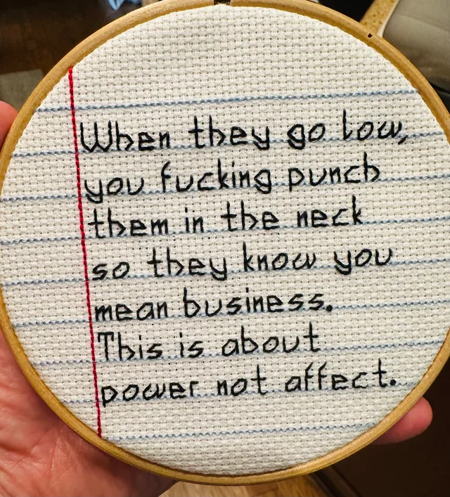 A cross-stitch in a wooden hoop, stitched to look like lined notebook paper with a red margin line and blue rules. In black block lettering it reads: "When they go low, you fucking punch them in the neck so they know you mean business. This is about power not affect."