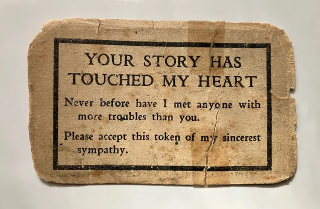 A worn, taped, cracked piece of old brownish card with a thick black rectangular border. Printed text reads: "YOUR STORY HAS TOUCHED MY HEART. Never before have I met anyone with more troubles than you. Please accept this token of my sincere sympathy."