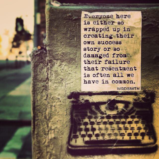 A weathered paper poster pasted on a gritty wall above a black stenciled typewriter. Typewriter-font text reads: "Everyone here is either so wrapped up in creating their own success story or so damaged from their failure that resentment is often all we have in common." Signed WRDSMITH.