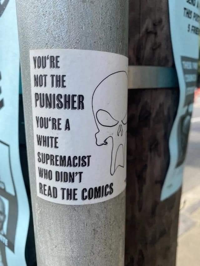A white sticker on a metal pole shows a black-and-white outline of a skull. Text reads: "YOU'RE NOT THE PUNISHER. YOU'RE A WHITE SUPREMACIST WHO DIDN'T READ THE COMICS". Blue flyers are on the pole and the wooden pole beside it.
